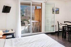 Ett hotellrum med en säng, ett skrivbord med en lampa, en TV, en balkong med stolar och en matplats med ett bord och stolar.