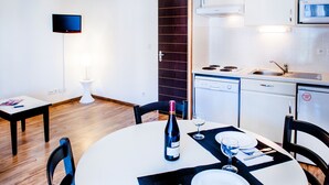 A dining area with a round table set for two, a bottle of wine, and a glass. There is a white table lamp, a small white pedestal table, and a white chair. The room has wooden flooring and a dark wooden door. - Residence Foch (Lourdes)