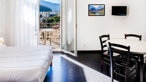 A hotel room with a bed, a dining table with chairs, a TV, and a balcony with a view of buildings and mountains. - Residence Foch (Lourdes)