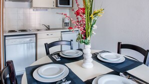 A dining area with a round table set for four, a white vase with flowers, and a kitchen in the background. - Residence Foch (Lourdes)