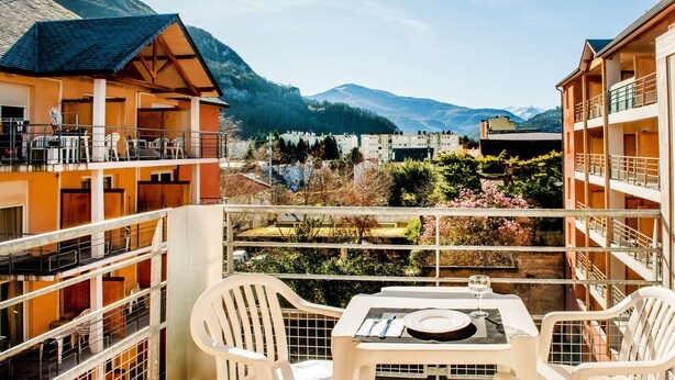 A balcony with white plastic chairs and a table set for two, overlooking a mountainous landscape with buildings in the background. - Residence Foch (Lourdes)
