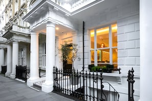 A white building with a black iron fence, columns, and a window with a view of a room. - The Porchester Hotel (London)