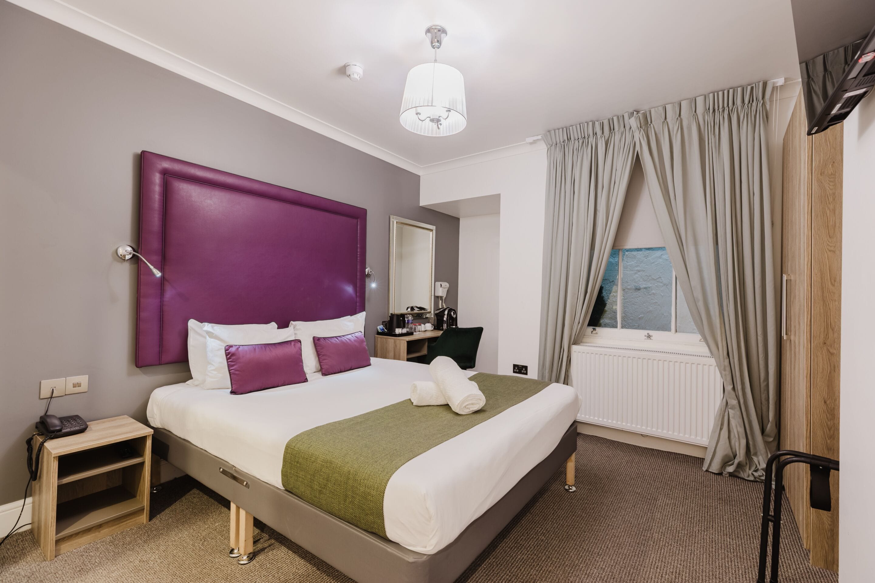 Une chambre d’hôtel avec un grand lit, un bureau, une chaise, un téléphone et une fenêtre avec des rideaux.