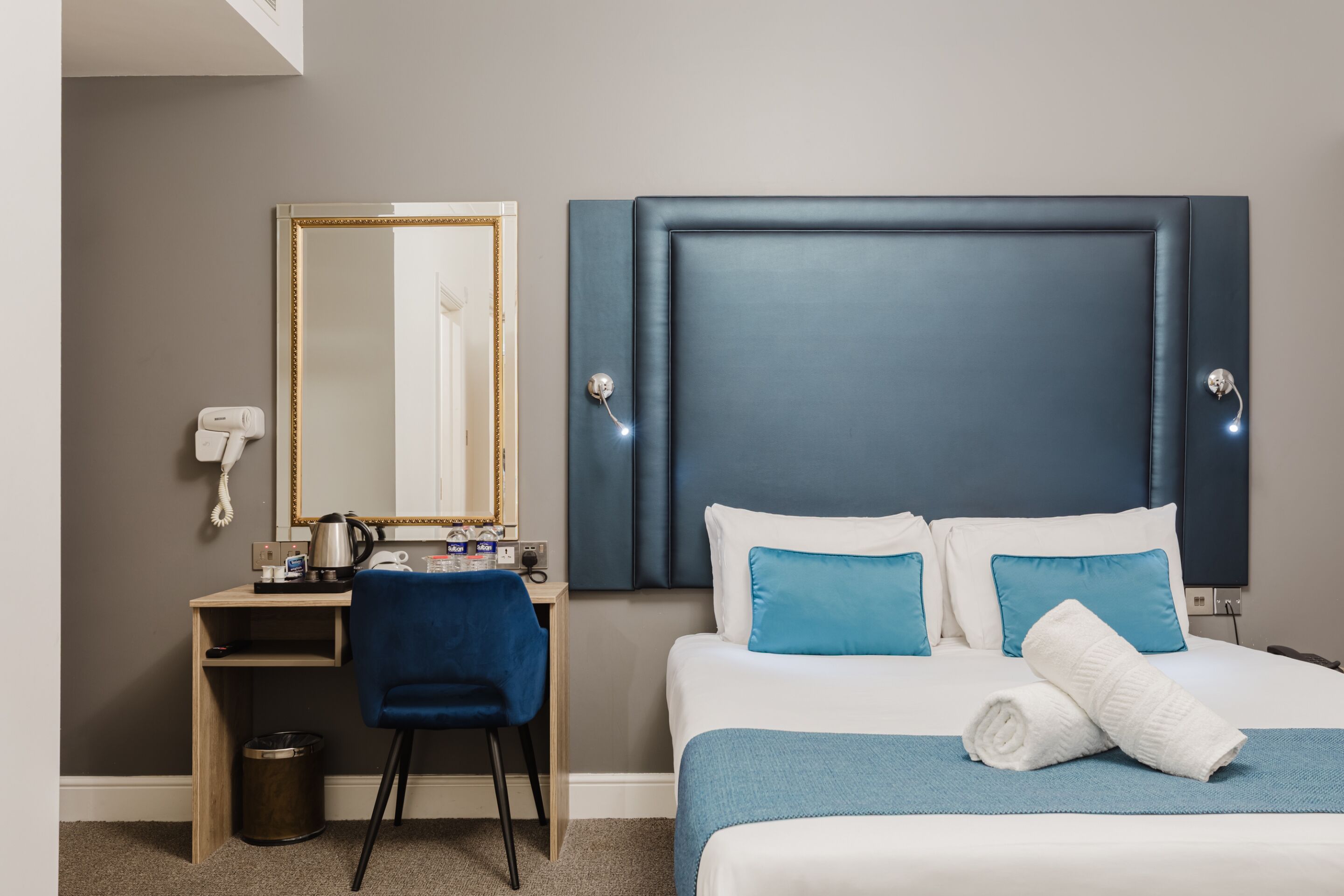A hotel room with a bed, a desk, a chair, a mirror, and a wall-mounted phone.