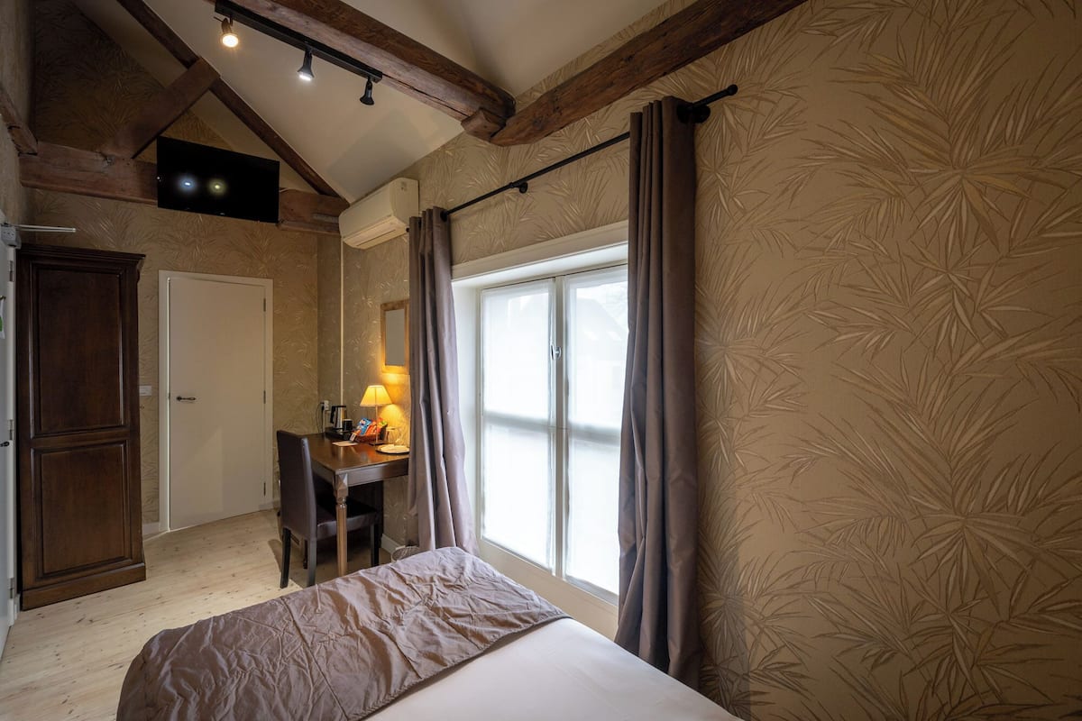 Un dormitorio con cama, escritorio con lámpara, armario y ventana grande con cortinas.