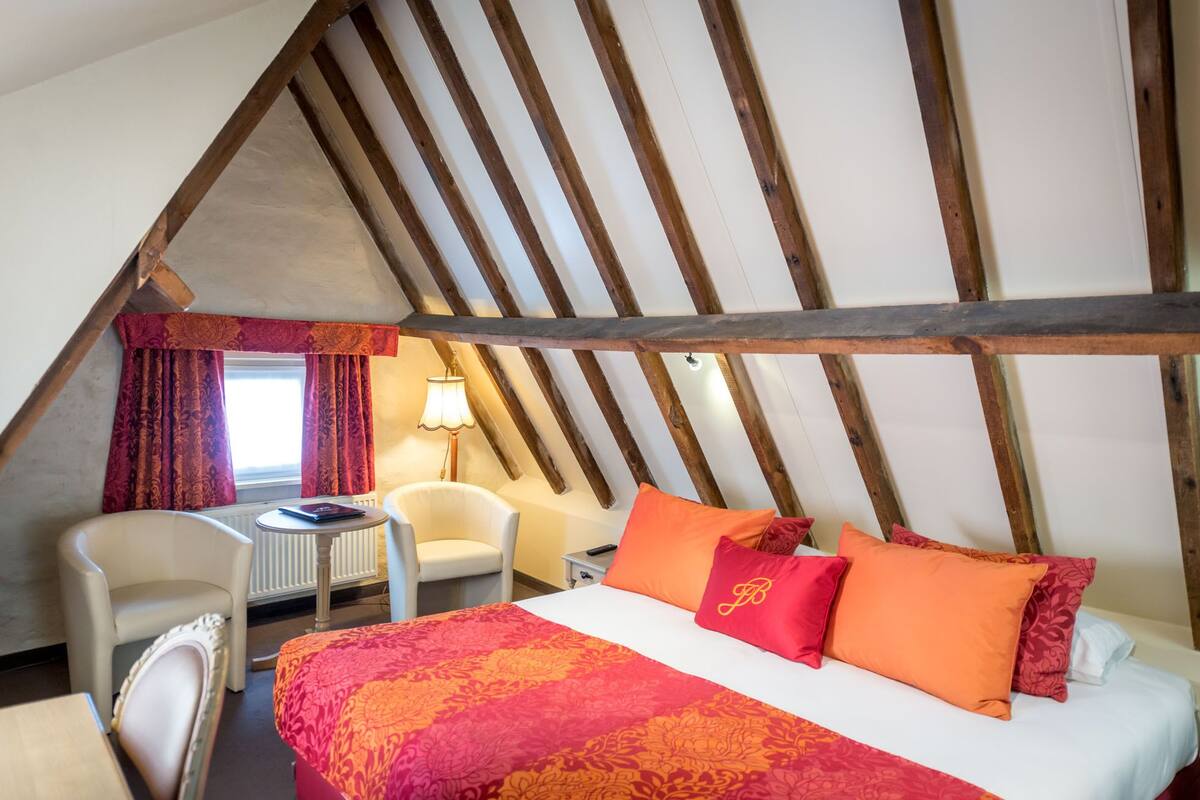 Un dormitorio con techo inclinado, una cama con ropa de cama roja y naranja, una silla blanca y una mesita de noche con una lámpara.