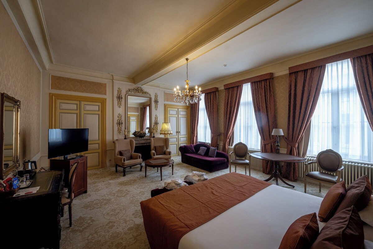A hotel room with a large bed, a TV, a desk, a sofa, and a chandelier.