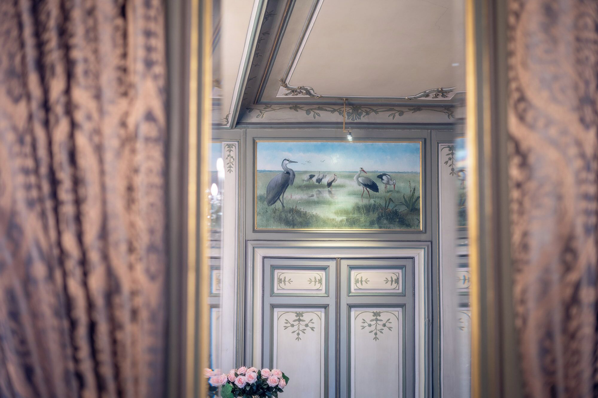 A room with a framed painting of birds on the wall, a mirror reflecting the room, and a vase of pink flowers on a table.