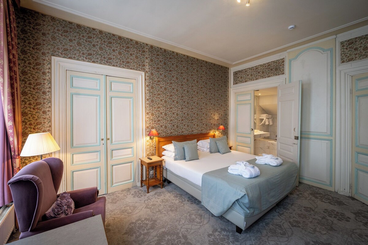A hotel room with a bed, two bedside tables, a chair, a bathroom with a shower, and a wardrobe.