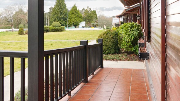 Une véranda couverte avec un sol carrelé, une balustrade en bois et une vue sur une zone herbeuse et des arbres en arrière-plan.