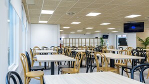 Une cafétéria avec de nombreuses tables et chaises, un téléviseur fixé au mur et une fenêtre laissant entrer la lumière naturelle.