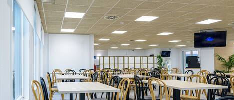 Una cafetería con varias mesas y sillas, un televisor montado en la pared y un ventanal que proporciona luz natural.