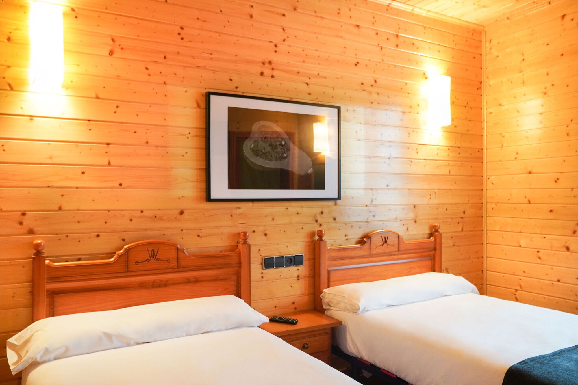 A wooden cabin room with two beds, a framed picture on the wall, and a nightstand with a lamp.