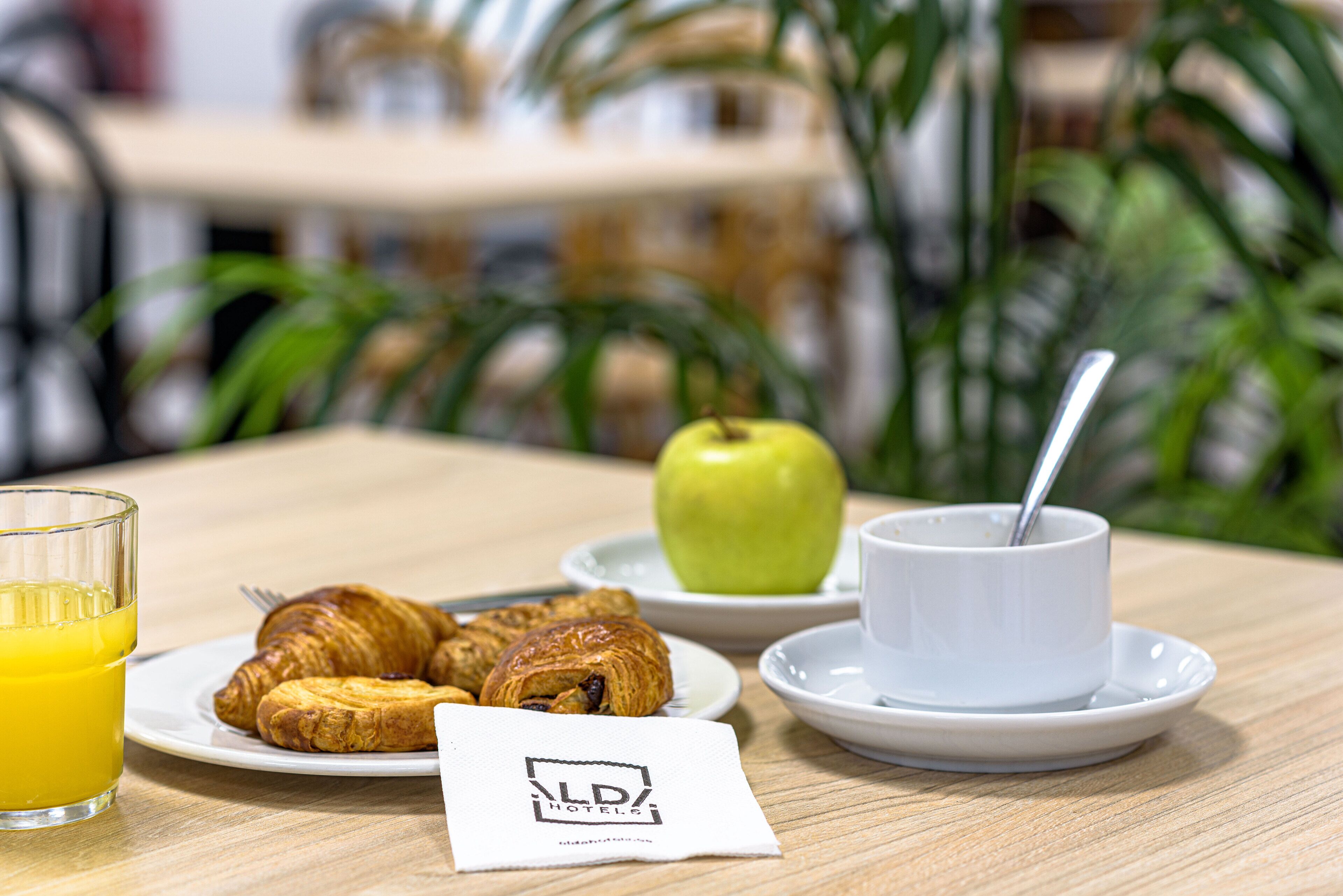 Ein Frühstückstisch mit Croissants, einem Apfel, einem Glas Orangensaft und einer Tasse mit Löffel.