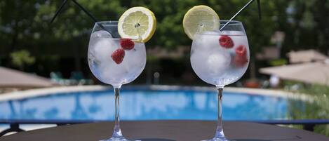 Deux verres de boissons glacées, décorés de tranches de citron et de framboises, sont posés sur une table au bord d’une piscine.