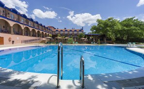 Una piscina con agua azul cristalina, rodeada de sillones de descanso y sombrillas, con un edificio de fondo que presenta arcos y detalles azules.