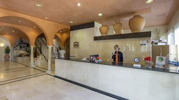 A hotel reception area with a marble counter, a receptionist, and a staircase with arches.