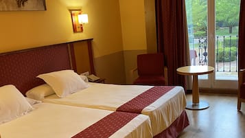 Een hotelkamer met twee bedden, een tafeltje, een stoel en een balkon met uitzicht op groen.