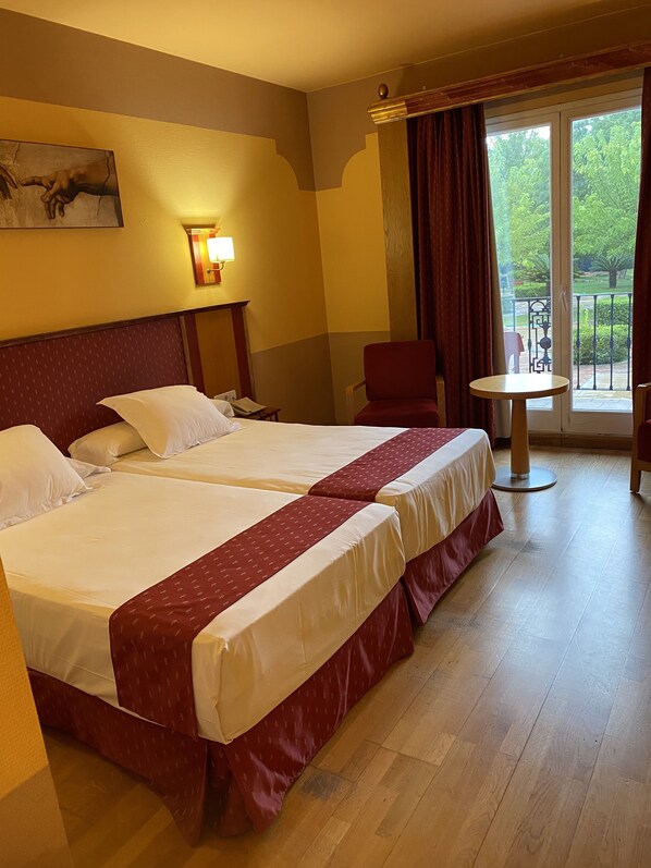 A hotel room with two beds, a small table, a chair, and a balcony with a view of greenery.