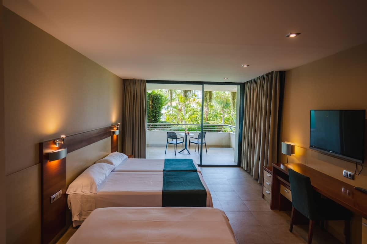 A hotel room with two beds, a desk, a TV, and a balcony with a table and chairs.