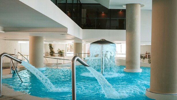 Una piscina interior con múltiples elementos acuáticos, incluyendo una cascada central y dos fuentes laterales.