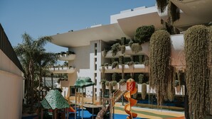 Zona infantil con toboganes y estructuras para escalar, rodeada de abundante vegetación y arquitectura moderna.