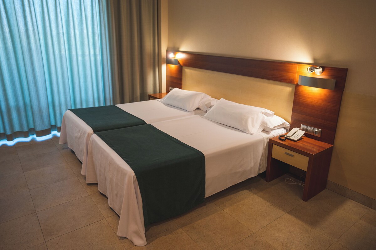 Una cama doble con cabecera de madera, dos mesitas de noche con lámparas, un teléfono y una ventana con cortinas.