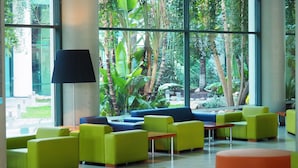 Un vestíbulo moderno con asientos verdes y azules, ventanas amplias y vistas a plantas tropicales.