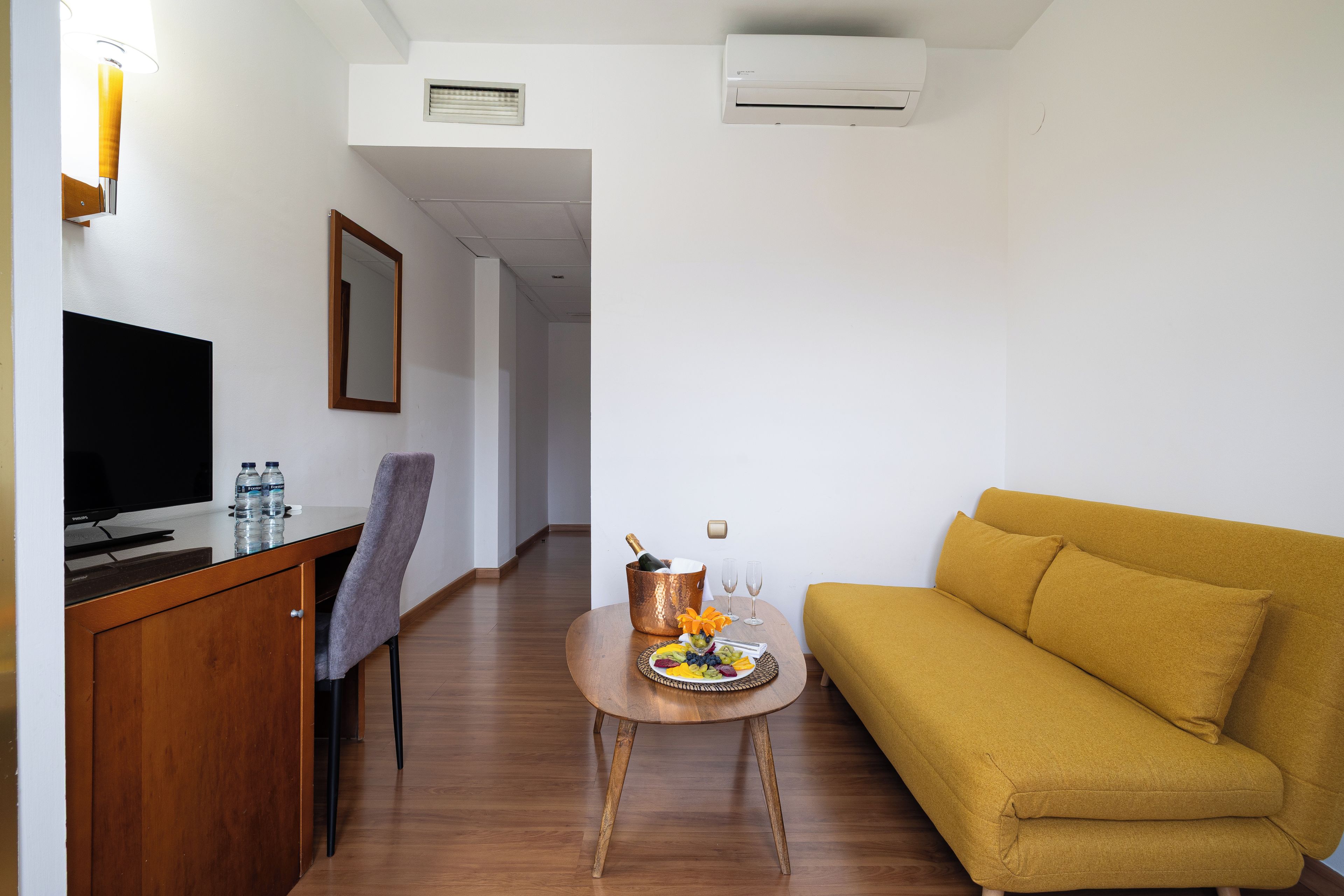 Habitación de hotel con un escritorio de madera, un televisor de pantalla plana, un sofá, una mesita con frutas y bebidas, y un espejo en la pared.