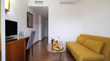 Habitación de hotel con un escritorio de madera, un televisor de pantalla plana, un sofá, una mesita con frutas y bebidas, y un espejo en la pared.