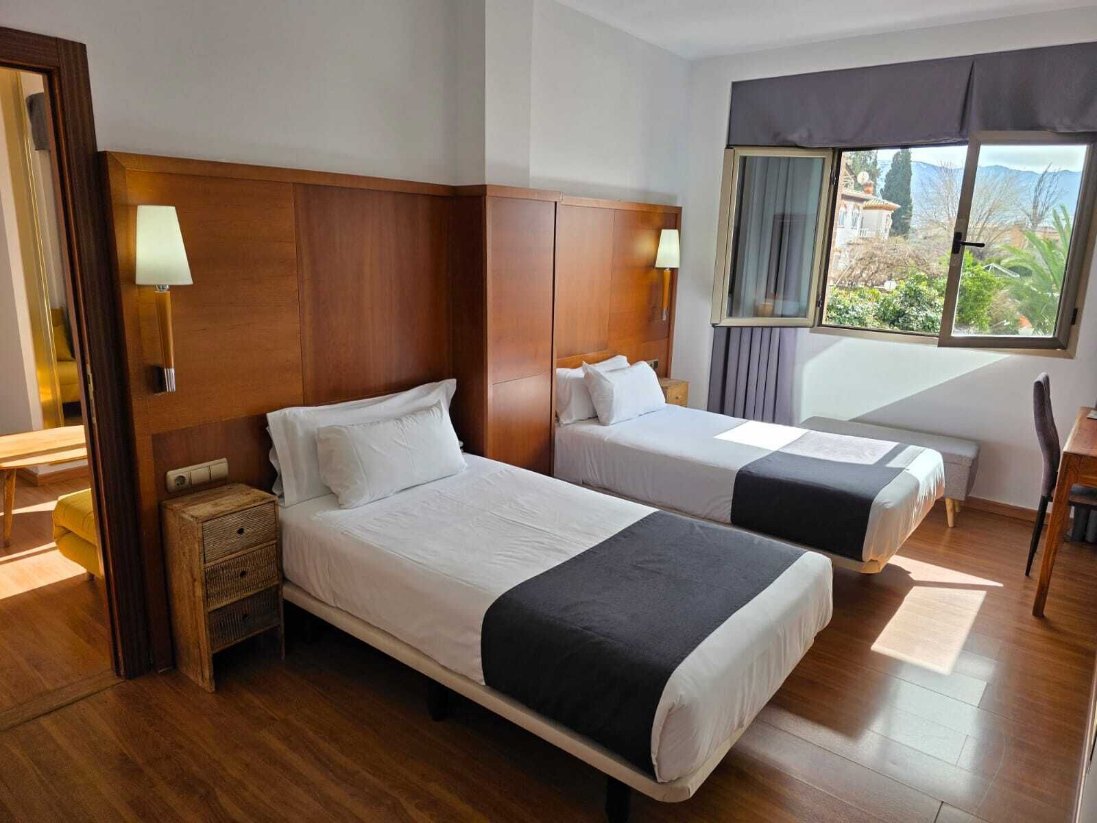 Habitación de hotel con dos camas, cabeceros de madera, una mesita de noche con lámpara, una ventana con vistas a la vegetación y una silla.