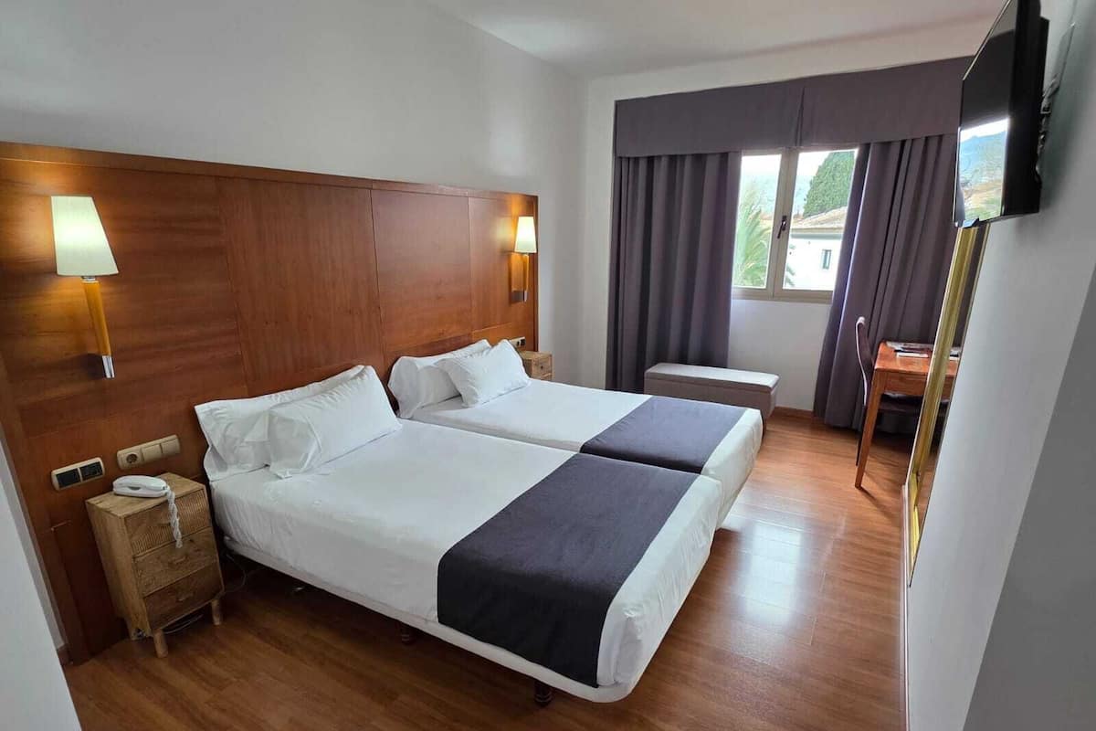 A hotel room with two beds, a wooden headboard, bedside lamps, a small bedside table, a TV, and a window with curtains.