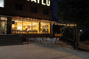 A building with a prominent illuminated sign, outdoor seating area with tables and chairs, and a staircase leading to the entrance. - Hotel Saylu (Granada)
