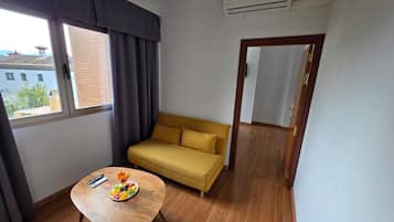 A room with a sofa, a table with fruit, a window with curtains, and an air conditioning unit.