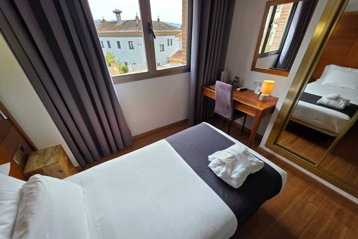 A hotel room with a bed, a desk, a chair, a lamp, and a view of buildings outside the window.