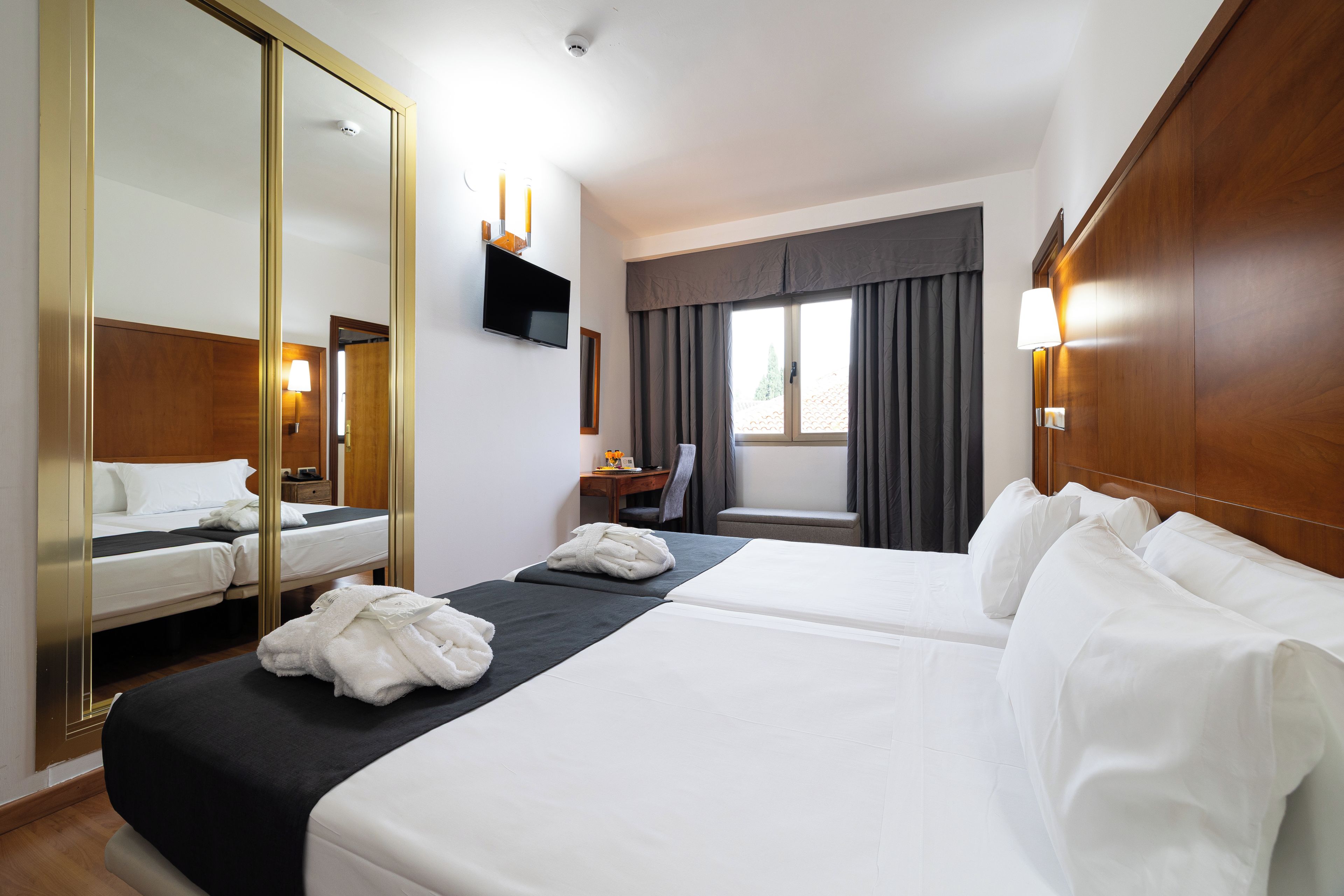 A hotel room with two beds, a large mirror, a TV, a desk with a chair, and a window with curtains.