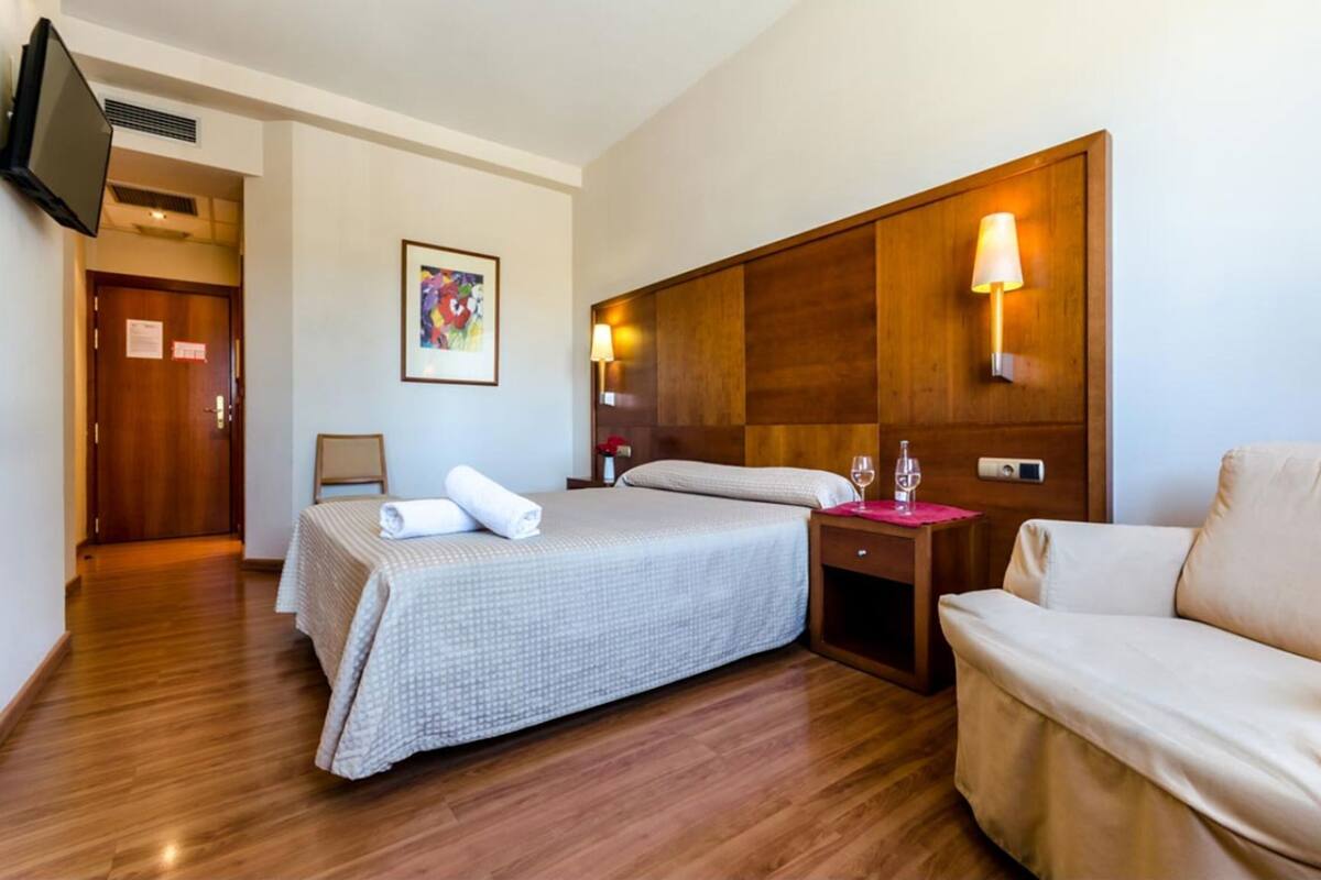 A hotel room with a bed, a sofa, a TV mounted on the wall, and a painting on the wall.