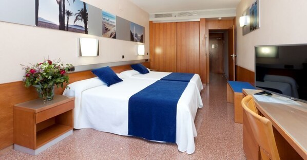 Habitación de hotel con dos camas, un escritorio con silla, un televisor y una pequeña mesa con un florero.
