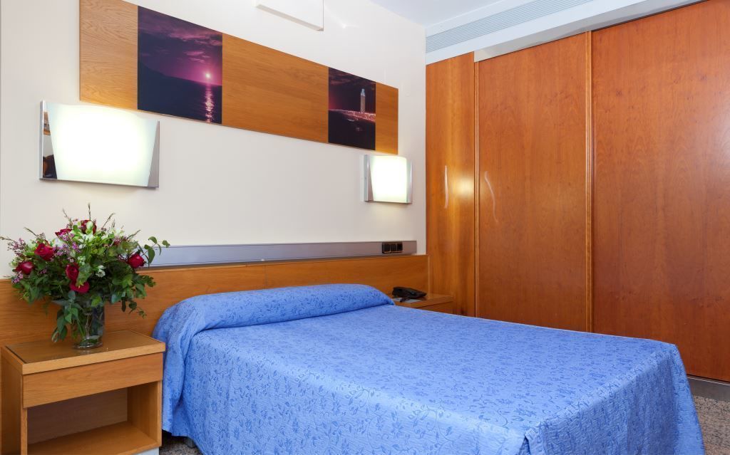 Una habitación de hotel con cama, mesita de noche, un jarrón con flores, armario de madera e iluminación empotrada.