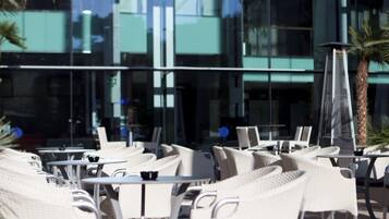 Una terraza con sillones y mesas de mimbre blanco, una palmera y un edificio moderno con ventanas amplias al fondo.