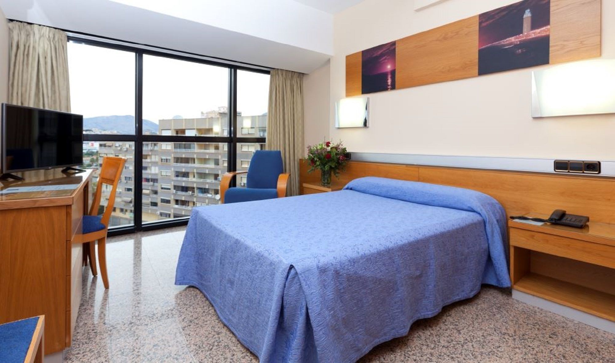 Habitación de hotel con cama, escritorio con televisor, una silla y un ventanal con vistas a la ciudad.