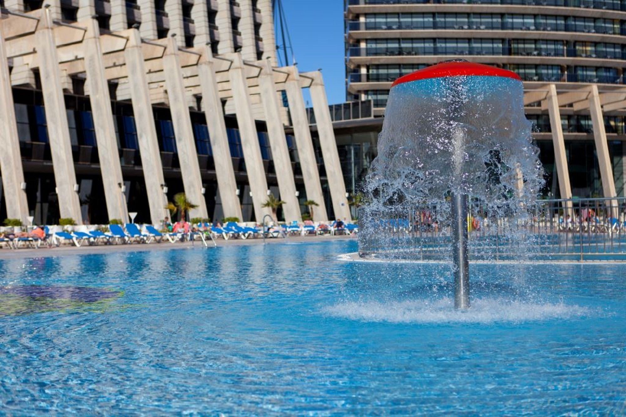 Una piscina con una fuente de agua en el centro, rodeada de sillas de descanso y un edificio moderno al fondo.