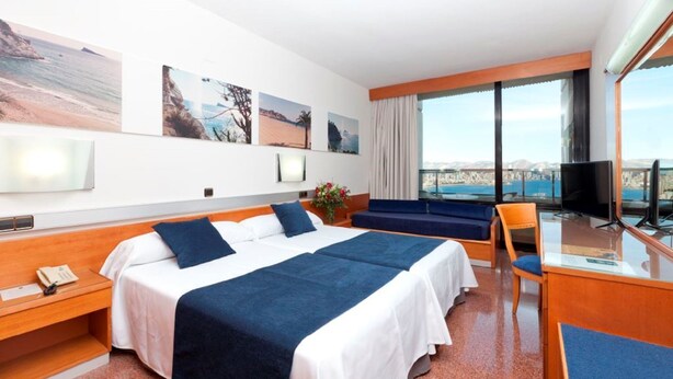 A hotel room with a large bed, a desk with a chair, a TV, and a view of the sea. - Gran Hotel Bali (Benidorm)