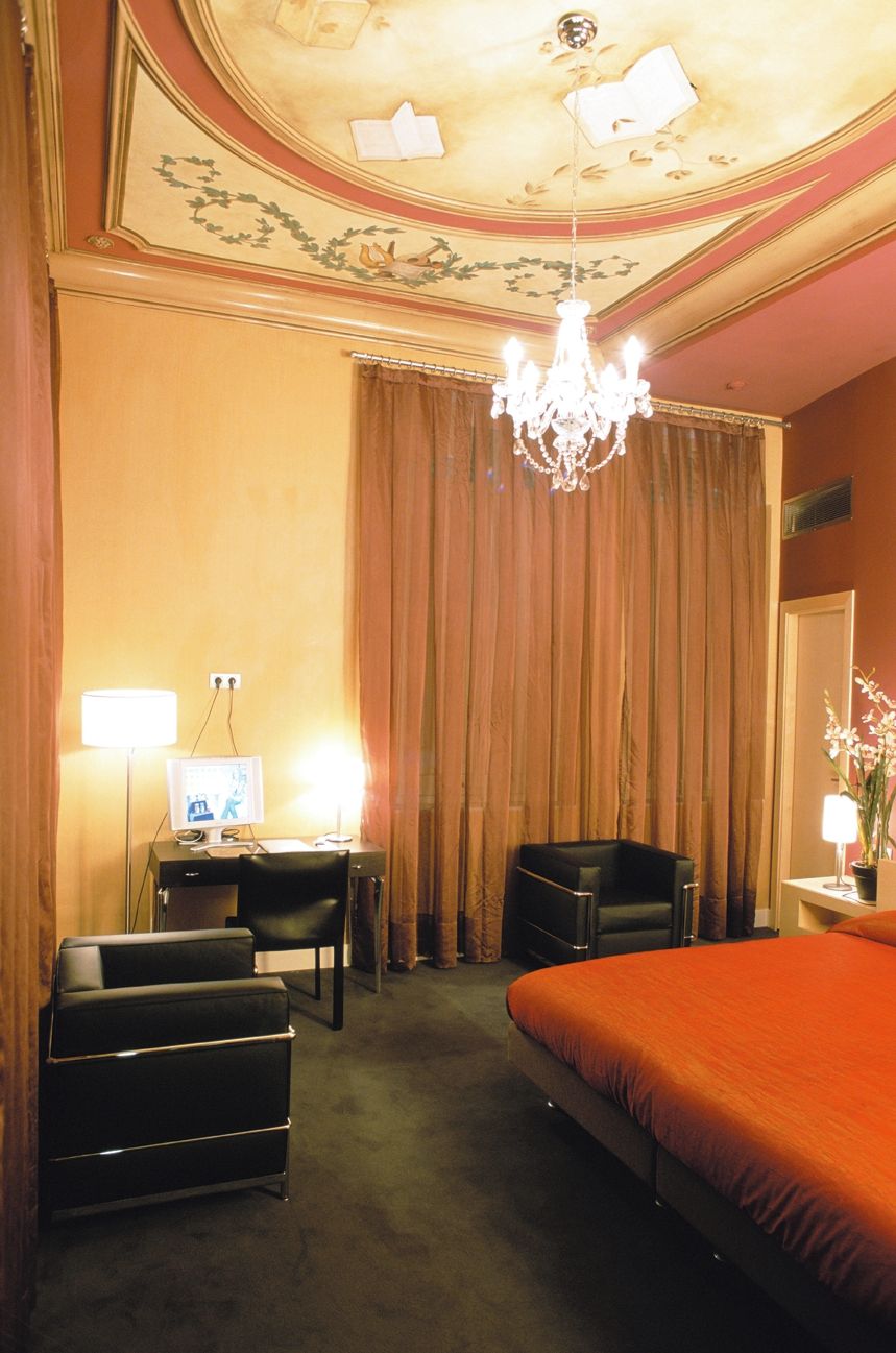 Habitación de hotel con cama, escritorio con computadora, silla, mesita con lámpara y candelabro.
