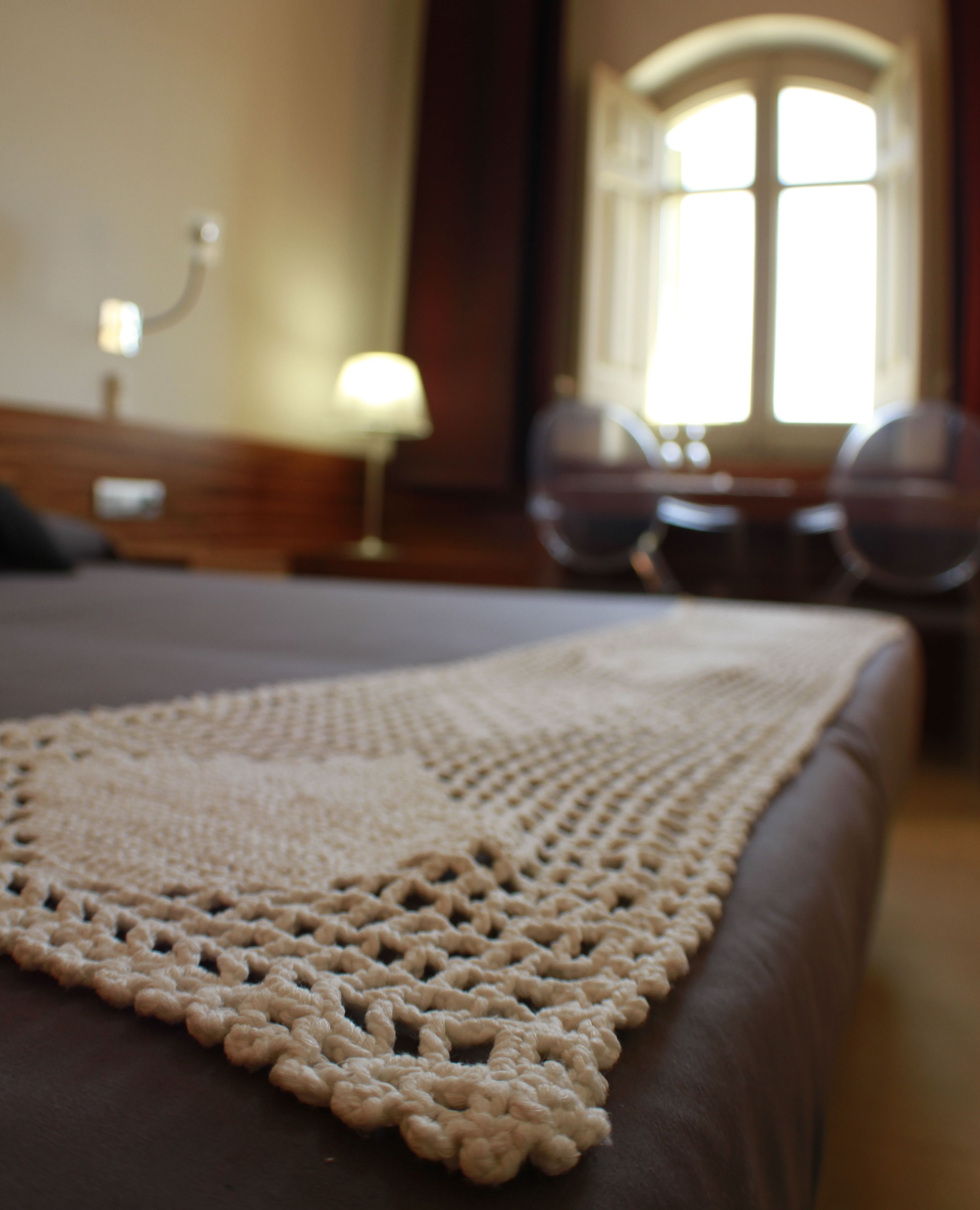 Una cama con un plaid de ganchillo, una mesita de noche con lámpara y una ventana con cortinas.