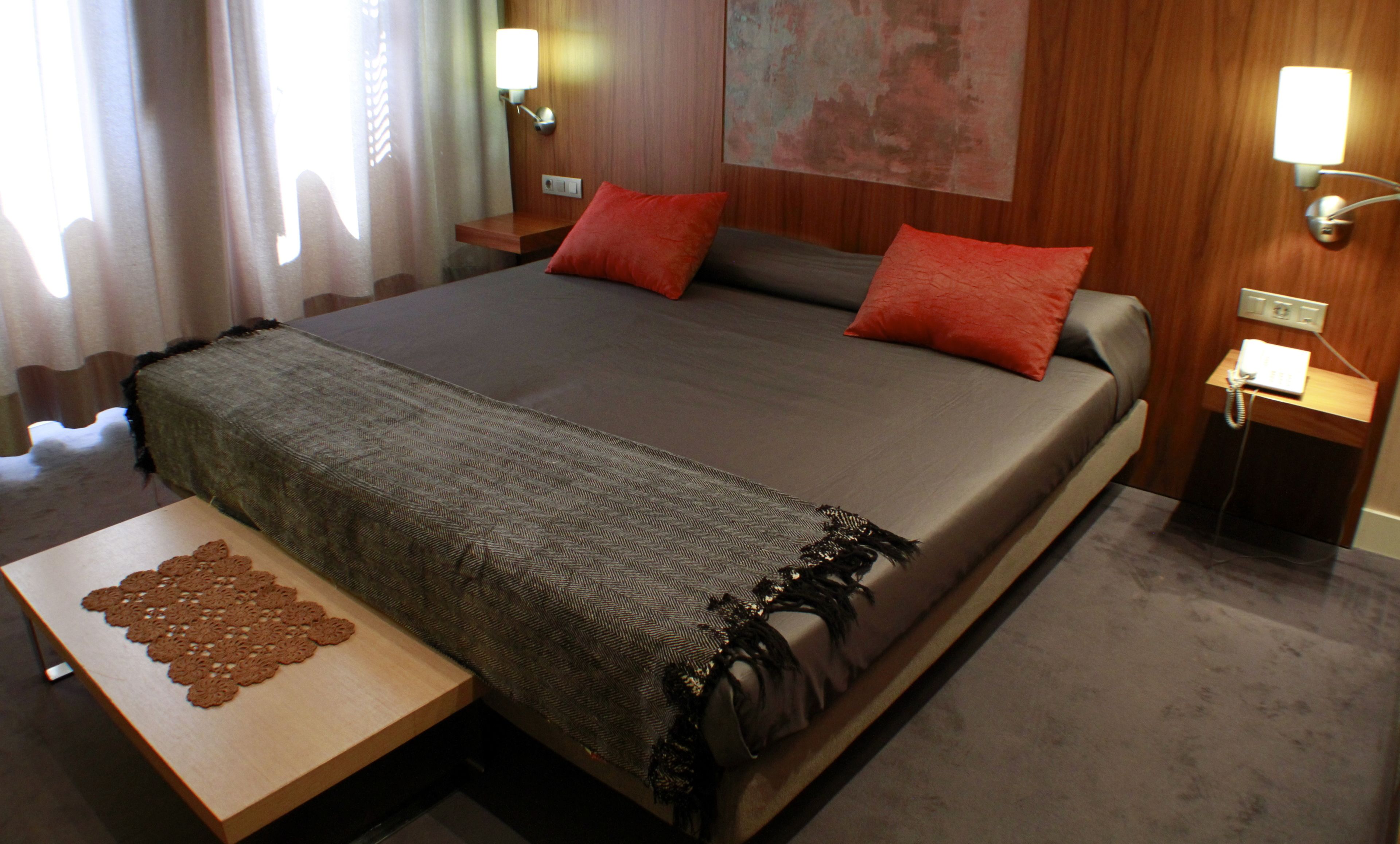 Habitación de hotel con una cama grande, dos mesitas de noche, una pequeña mesa con un mantel de encaje y un teléfono en la pared.