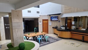 A hotel lobby with a reception desk, a wooden table with chairs, a small garden area with artificial grass and a potted plant, and a television mounted on the wall. - Ejido Hotel (El Ejido)