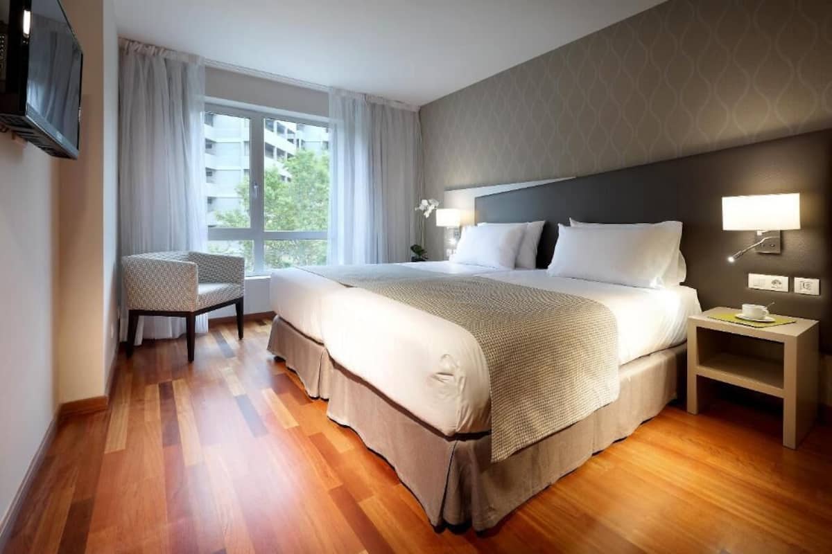 Habitación de hotel con una cama grande, una silla, un televisor, una mesita de noche con lámpara y una ventana con cortinas.