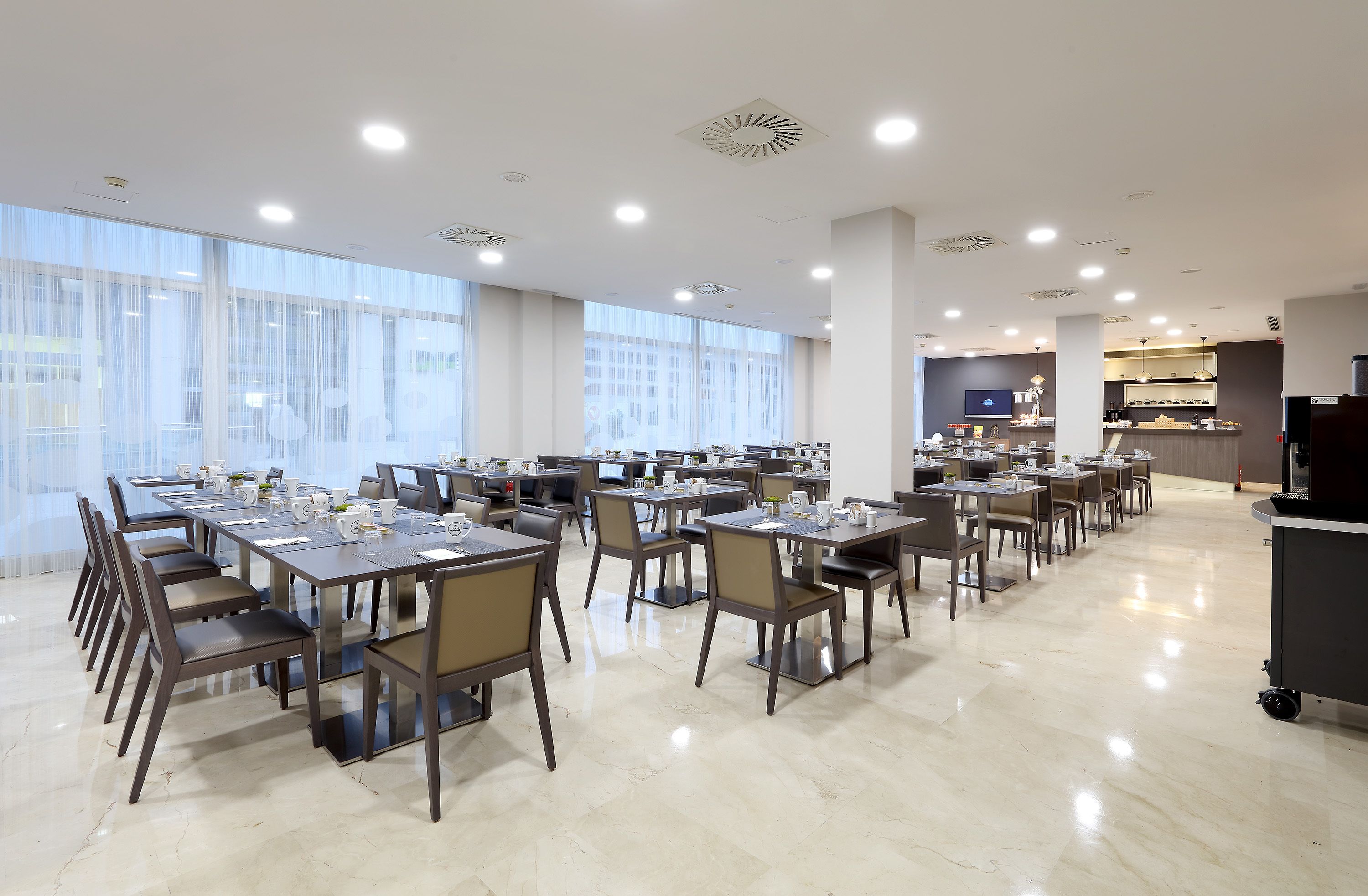 A spacious dining area with multiple tables and chairs, large windows, and a kitchen area in the background.