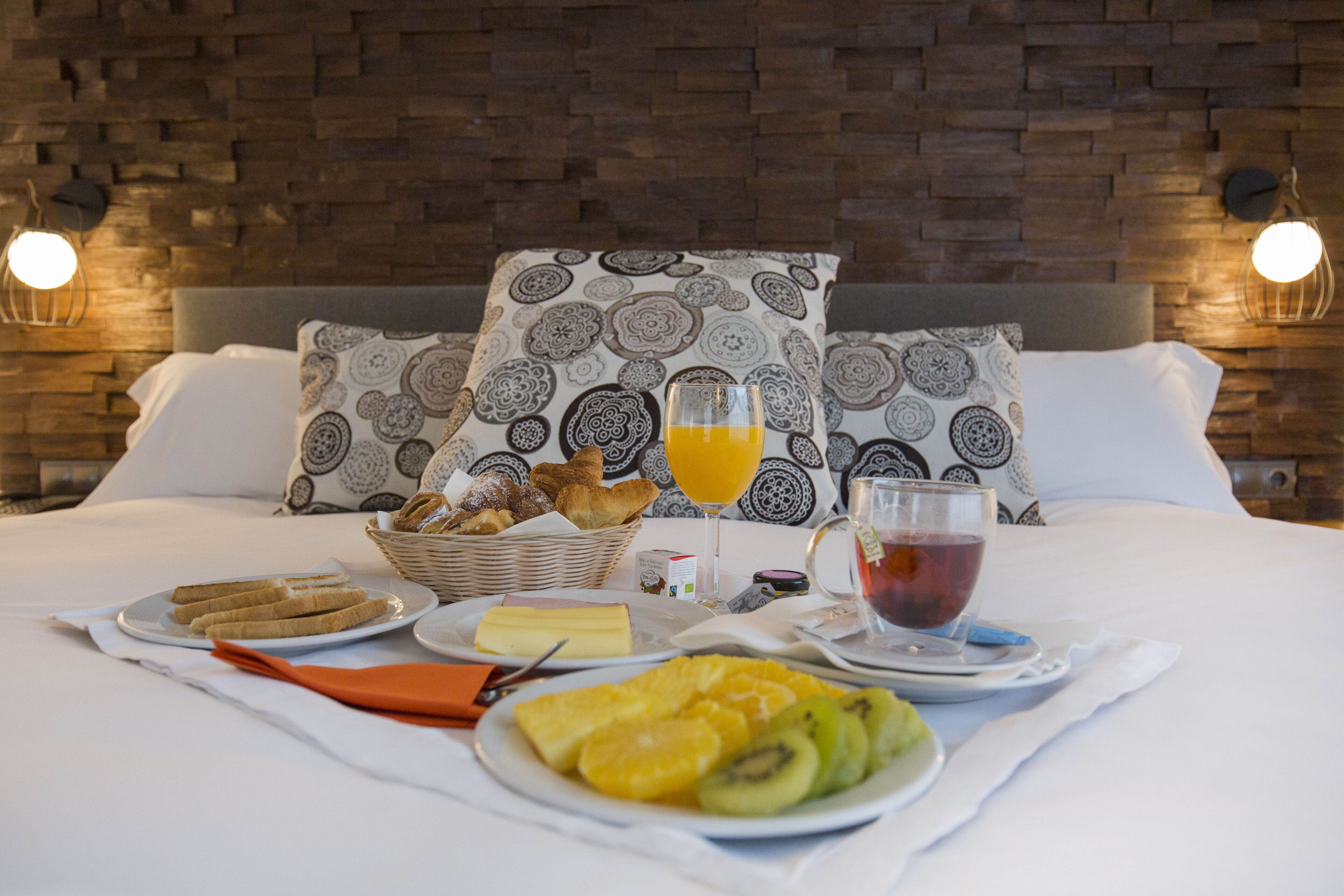 A neatly arranged breakfast table with plates of fruit, bread, and beverages on a bed with patterned pillows and a brick wall background.